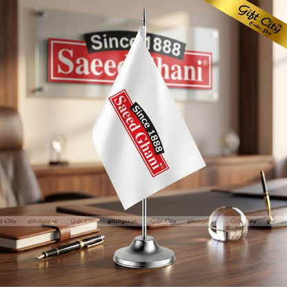 A small white desk flag on a silver stand, printed with the Saeed Chaini logo, displayed on a wooden desk in an office setting with a blurred Saeed Chaini sign in the background.