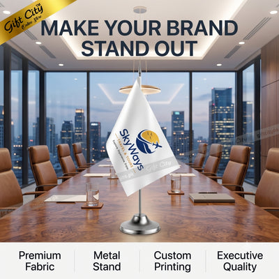 A customized desk flag on a silver metal stand displayed in a modern conference room with a city skyline view. The flag is white with a blue and yellow logo that reads 'SkyWays'.