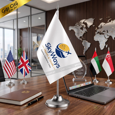 A white Skyways desk flag on a silver metal stand, displayed on a wooden conference table alongside other miniature national flags, a laptop, and a notebook in a modern office setting.